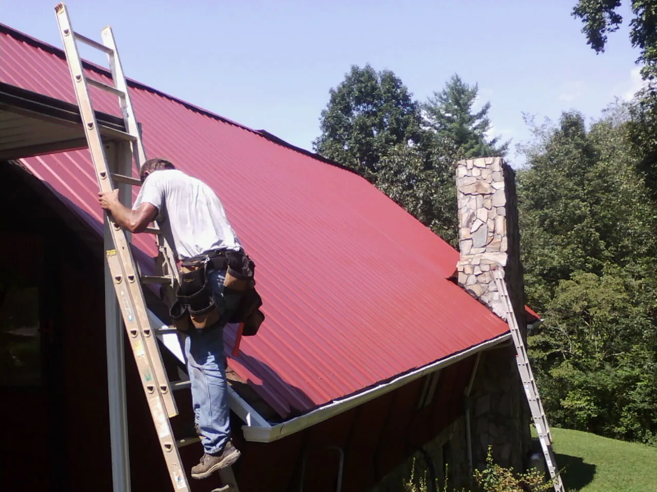 Roofer on a ladder beside a red metal roof and stone chimney — Soffit & Fascia Repair by Roofing Brothers in Bozman